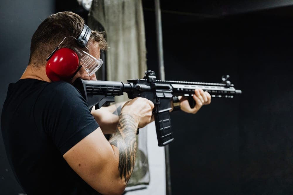 A shooter in red earmuffs and clear safety glasses aims a rifle at an indoor range