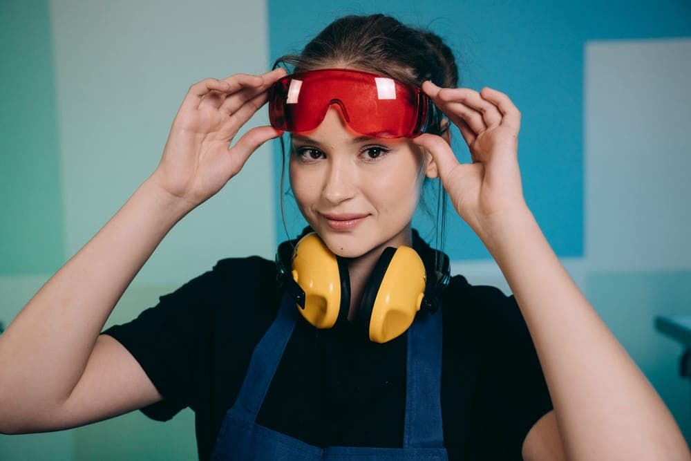 A smiling woman adjusting red safety glasses with yellow earmuffs around her neck