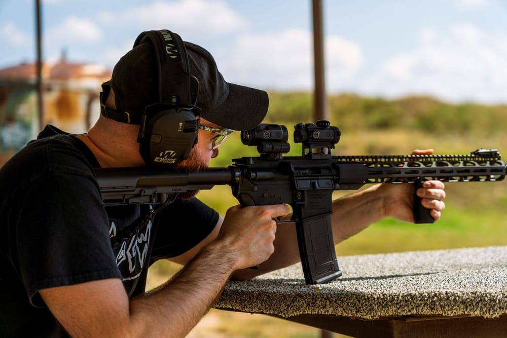 A shooter wearing electronic earmuffs and glasses aims from a bench at an outdoor range
