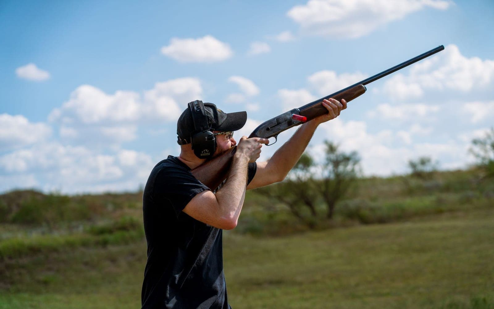 A shooter wearing ear protection and glasses aims a shotgun skyward at an outdoor range